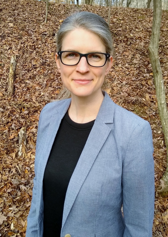 Photo of Havala Pye in the woods wearing a grey blazer and black glasses