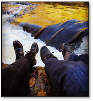 Feet pointing toward a small waterfall, taken from the point of view of the photographer.