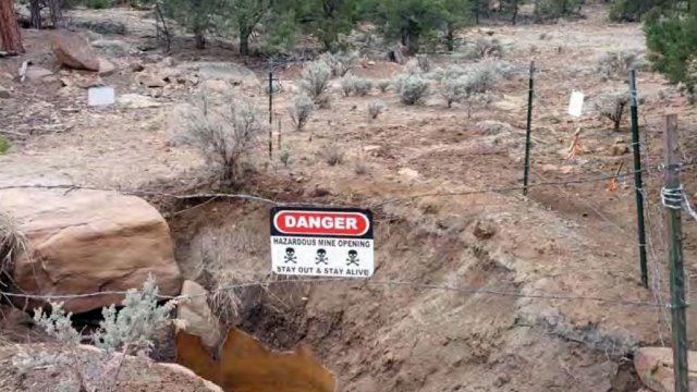 sign attached to barbed wire surrounding a hole in the ground reads: DANGER: HAZARDOUS MINE OPENING - STAY OUT & STAY ALIVE"