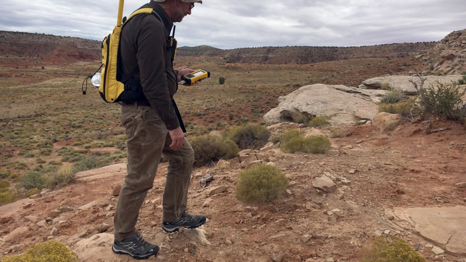 Researcher is walking though desert region with a backpack mounted GPS locator and a handheld device to scan for gamma radiation