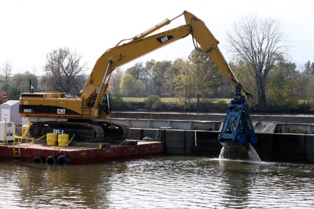 Workers use excavators with environmental clamshell buckets mounted on flat, anchored platforms to dredge the river. The PCB-contaminated sediment is emptied onto 35-foot-wide, 195-foot-long floating barges.