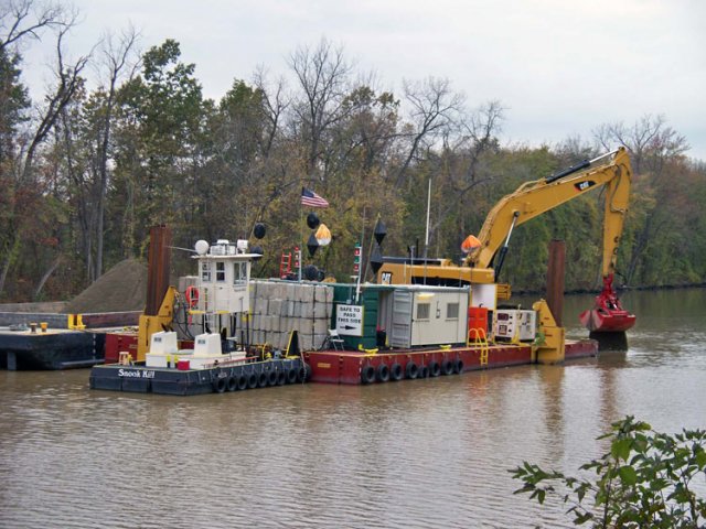 Workers use excavators with environmental clamshell buckets mounted on flat, anchored platforms to place backfill on top of the previously dredged area, returning the riverbed to its natural state.