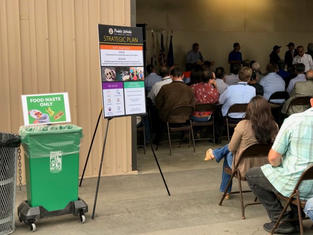 This is a meeting with rows of people attending with a food waste bin next to a Public Works strategy sign.
