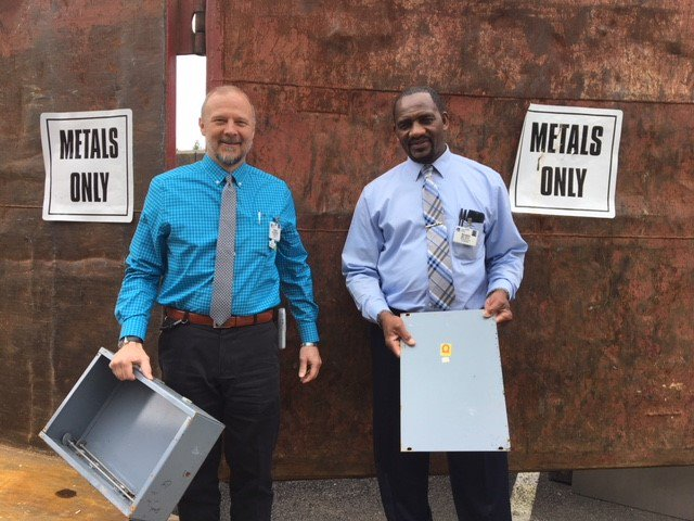 This is a picture of two men holding items in front of a bin that has two signs that read "METALS ONLY."