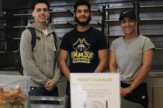 This is a photo of three UMASS Dartmouth students posing in front of a "Project Clean Plate" poster. Project Clean Plate is UMASS Dartmouth food recovery initiative.