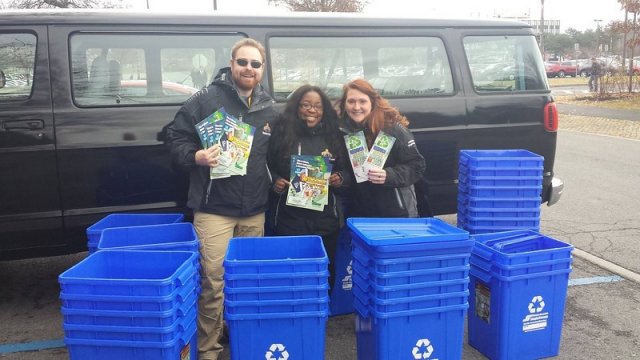 This is a picture of three students from University of Albany standing next to numerous blue recycling bins.