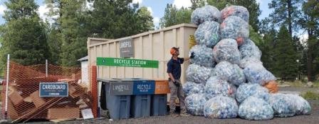Worker standing next to dumpster with recycling bins and tall stack of bagged waste