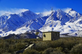 Small building with radio antenna on valley floor with snowcapped peaks in background.