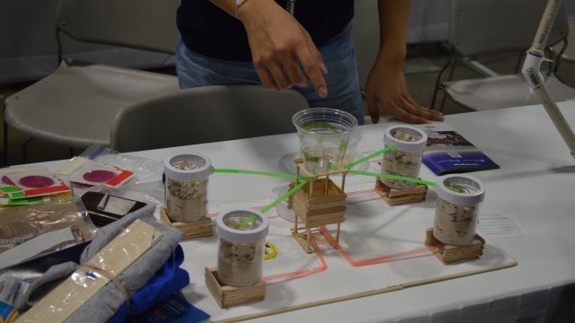 Closeup of the UCDavis Greywater project: a plastic cup sitting in a popsicle stick structure and connected with straws to cylinders