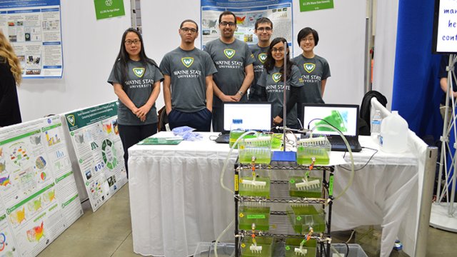 Six Wayne State people smiling and posing with their tanks of algae water in front