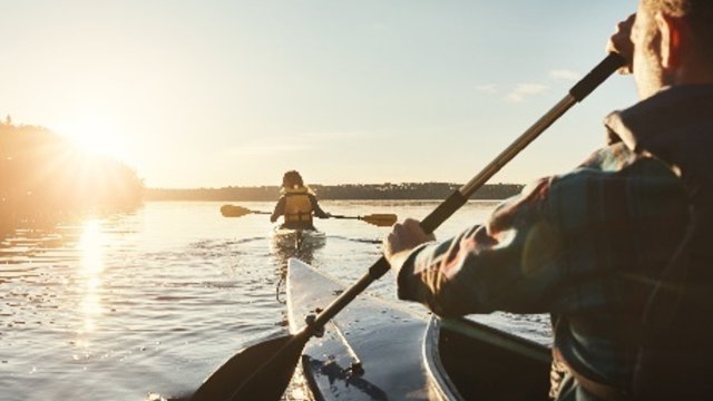 A person paddles a kayak in a calm body of water, while facing into a sunset
