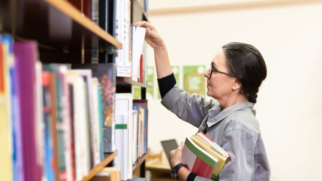 A librarian holding books reaches for one on a library shelf.