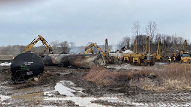 Burned train boxcars being moved by large excavation equipment