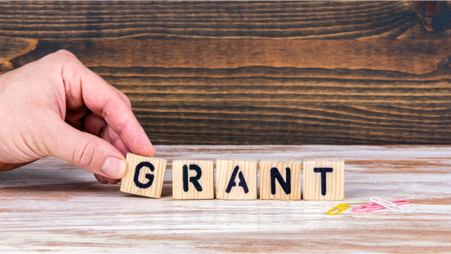 A hand is placing the letter G in a row of wooden block letters spelling the word grant. Three is a brown wood background. 
