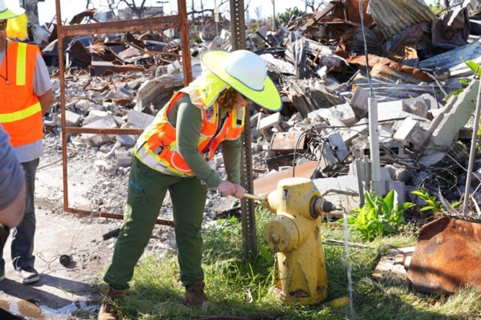 An EPA worker in safety gear is opening a fire hydrant. Behind the worker is a destroyed building.