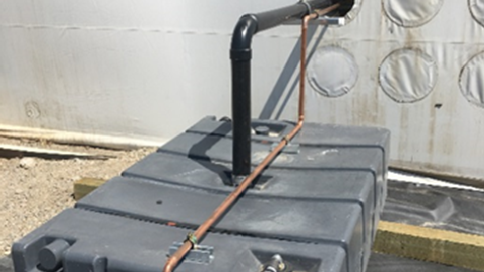 A black box connected with various water pipes, two of which are entering a white building behind the box.