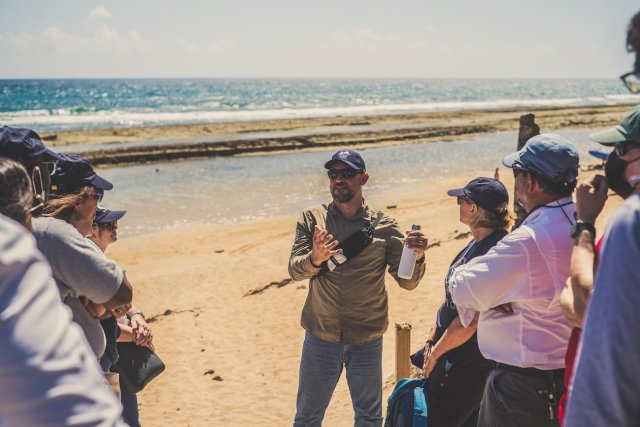 A group gathered around a professor at the beach, detailing plans for dune restoration to support coastal resilience.