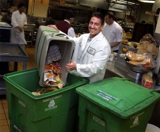 Restaurant employee pours food scraps into a large green compost bin in a commercial kitchen.