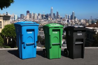 Blue recycling, green compost, and black trash collection bins on a San Francisco street with San Francisco skyline in the background.