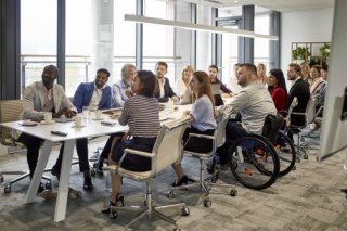 A group of roughly a dozen business people sit around a conference table, including one man who uses a wheelchair.