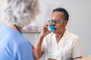 Older woman using an inhaler while a nurse (with back turned to camera) looks on.