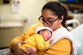 Mother wearing a yellow shirt holds a newborn with a yellow jumper while resting on a hospital bed.