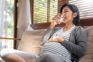 A pregnant mother sits on a couch drinking a glass of water.