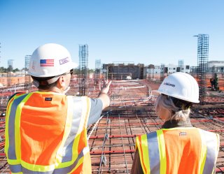 Two workers with hard hats and vests overlook a construction site in process.