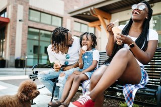 A young, Black family of three sits on a bench and eats ice cream. Their small dog watches them.