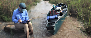 Researcher writing notes in a stream alongside a paddle boat with research equipment on it