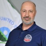 A light-skinned man with a bald head and a salt-and-pepper goatee. He is wearing a dark blue EPA polo shirt and stands in front of the EPA flag.