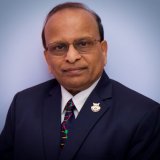 A brown-skinned man with glasses, in a dark blue suit.