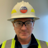 A light-skinned man wearing an EPA hard hat, safety glasses, and a high-visibility vest.
