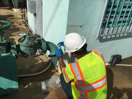 Worker in yellow safety vest and hard hat squatting down near pipes to perform water sampling.