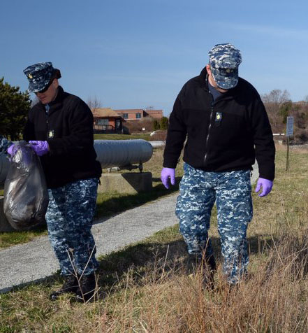 Two people wearing navy fatigues and gloves walking in grass next to a road, one is placing something in a garbage bag.