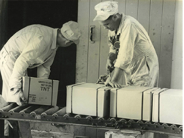 Historic photo of two people grabbing boxes of TNT from a conveyor belt