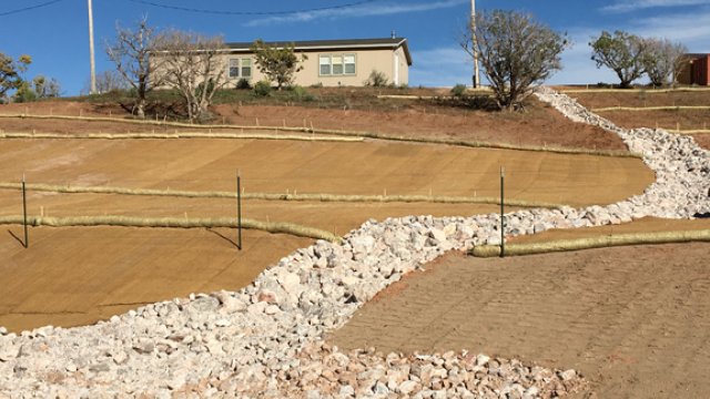 Steep slope no longer showing signs of water erosion. Instead berms in place and a large, stone filled channel for water goes up the slope leading to a home