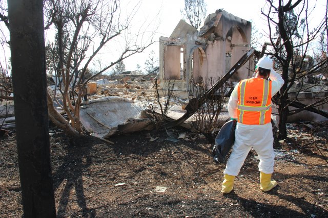 Worker in bright orange safety vest emblazoned EPA Contractor on back surveys a burned home