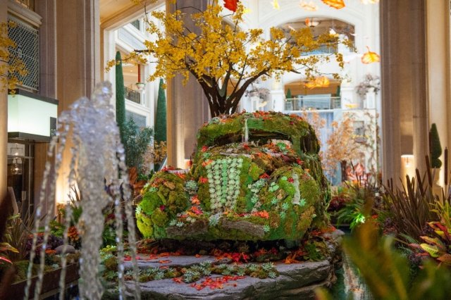 This is a picture of artwork that that resembles nature. There's a tree with yellow foliage in the background, a small water fountain in the foreground, plants all around, and an old-fashioned car covered with grass and leaves in the center.