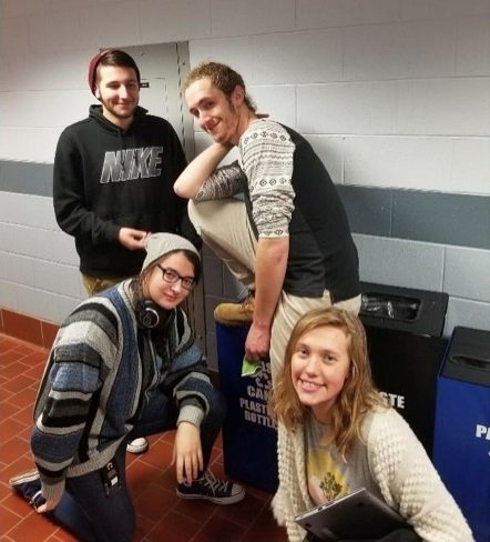 This is a picture of four Raritan Valley College students posing for the camera in front of various recycling bins inside a building.