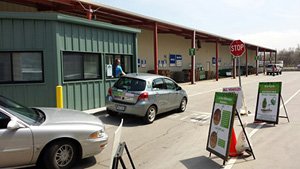 Cars lined up to pass by booth. Driver is speaking to booth attendant. Warehouse in the background.