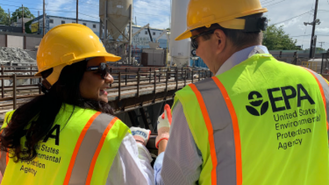 One male and one female EPA employees wearing yellow safety helmets and yellow vests with the EPA logo on the back.
