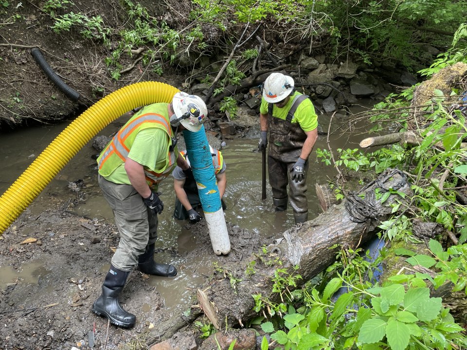 Two crew members use a large vacuum hose to capture oily sheen and sediment in the stream.