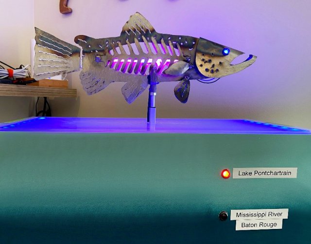 The kinetic fish sculpture uses colored light and motion to visualize Village Blue Lake Pontchartrain water quality data. The purple color of the sculpture, shown here, indicates high salinity levels; the blue color of the base represents low algal levels; and the low height of the fish corresponds to low turbidity levels. Data shown represent measurements taken from Lake Pontchartrain, noted by the red light on the front.