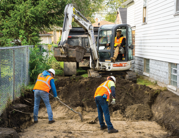 Photograph of two men digging a large hole in the ground next to a house. A mini excavator can be seen behind them assisting with the dig.