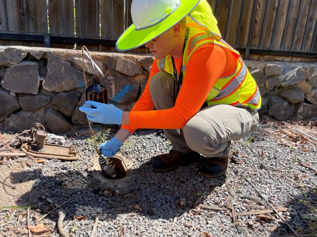 Worker in high visibility vest crouching next to a drinking water access panel taking a water sample.