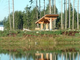 Landscape view of a modern wooden shelter on the banks of a placid lake. Coniferous trees are seen in the background.