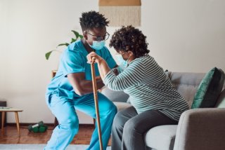 A nurse helps an older woman with a cane get up off of a couch.