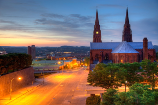 An aerial view of Albany, NY at dusk with orange glow of streetlights, steeples in the background against sky and trees in foreground. 