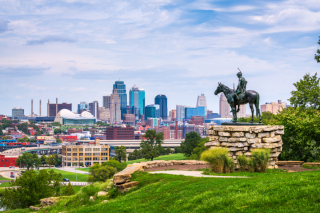 An elevated statue in the foreground of a horse and rider is surrounded by green grass and a stone wall. The background is the skyline of Kansas City, MO against a blue sky with white clouds. 